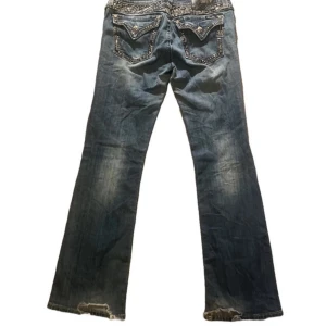 Blå jeans med detaljer - Säljer ett par blå miss me jeans med snygga slitningar och dekorativa detaljer på bakfickorna. Jeansen har en bootcut-stil och är i bra skick med lite slitage vid benslutet. strl 31 midja: 38 innerbenslängd: 85