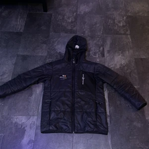 Svart pufferjacka från Sail Racing  - Säljer en svart pufferjacka från Sail Racing i mycket bra skick. Jackan har en dragkedja framtill och en huva för extra skydd mot väder och vind. Perfekt för kalla dagar med sin quiltade design. Den har också flera praktiska fickor med dragkedjor.