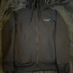 Grå hoodie från McKenzie - Säljer en grå hoodie från McKenzie i bra skick. Den har dragkedja och en klassisk huva med snören. Perfekt för höst och vinter, och funkar lika bra till vardags som till träning. Materialet är mjukt och bekvämt, vilket gör den till en favorit i garderoben. 🖤(Hör av dig om du vill ha med byxorna som finns till settet så löser jag 🤗)