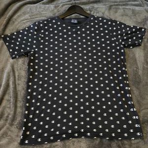 Svart t-shirt med logotypmönster från Champion - Säljer en svart t-shirt från Champion i storlek S. Den har ett coolt mönster med Champion-logotypen över hela tyget. Perfekt för en avslappnad stil eller träning. T-shirten är i mycket bra skick och gjord av mjukt material för extra komfort.