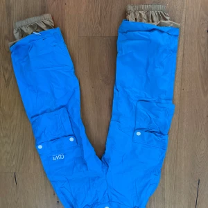 Blå skidbyxor från CLWR - Säljer ett par blå byxor från CLWR i storlek 140. De har praktiska fickor med knappar och elastiska muddar vid bensluten. Perfekta för utomhusaktiviteter under vinter och i backen. Byxorna är i bra skick och redo för nya äventyr!