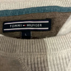 Grå tröja från Tommy Hilfiger - Säljer en stilren beige tröja från Tommy Hilfiger i storlek L. Tröjan är i bra skick och har en klassisk design med ribbade muddar vid ärmslut och nederkant. Perfekt för både vardag och lite finare tillfällen. Passar bra till jeans eller chinos!
