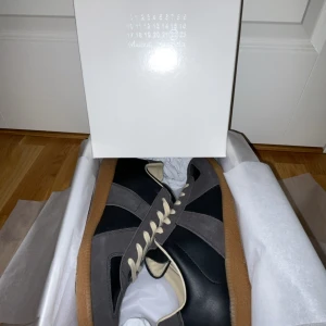Svarta och grå sneakers från Maison Margiela - Säljer ett par snygga sneakers från Maison Margiela i svart och grått. Helt nya med ett otroligt bra skick! Allt på bilderna med följer!