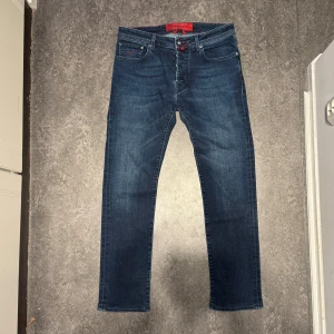 Mörkblå jeans från Jacob Cohen - Säljer ett par snygga mörkblå jeans från Jacob Cohen. De har en klassisk femficksdesign och röd detaljsömnad som ger en cool kontrast. Jeansen är i mycket bra skick och passar perfekt för både vardag och fest. Märkeslappen i rött på baksidan ger en extra touch av stil.