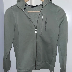 Olivgrön zip hoodie från Sail Racing - Säljer en snygg olivgrön zip hoodie från Sail Racing i storlek 150. Storleken stämmer inte skulle jag säga då den fortfarande passar mig som är 170. Tröjan har en dragkedja framtill och en luva. Perfekt för kyliga dagar och passar till både vardag och fritid. Hoodien är i mycket bra skick och redo för en ny ägare! Priset går att diskutera vid snabb affär.
