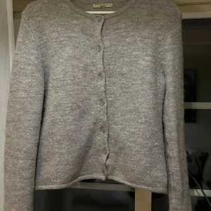 Grå kofta från Gina Tricot - Mysig grå kofta från Gina Tricot i storlek S. Den är hög kvalitet, stickad och har knappar hela vägen fram. Perfekt för kyliga höst- och vinterdagar. Koftan är i mycket bra skick men har ett litet hål vid höger arm, jättelätt att laga!. Passar både till vardags och lite finare tillfällen.