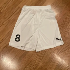 Vita shorts från Puma med nummer 8 - Säljer ett par vita shorts från Puma i mycket bra skick. De har en elastisk midja och är perfekta för fotboll eller träning. På ena benet finns en svart siffra 8 och Pumas logga. Materialet känns lätt och bekvämt, perfekt för sommarens aktiviteter. Funkar till både tjej och kille.