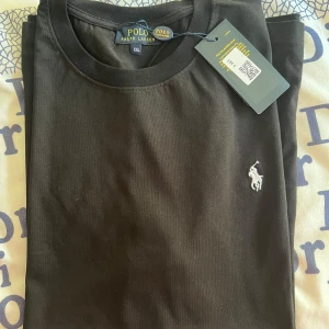Svart t-shirt från Polo Ralph Lauren - Säljer en snygg svart t-shirt från Polo Ralph Lauren i storlek XL. Den är i nytt skick med etikett kvar. T-shirten har en klassisk design med en liten broderad logga på bröstet. Perfekt för både vardag och fest!
