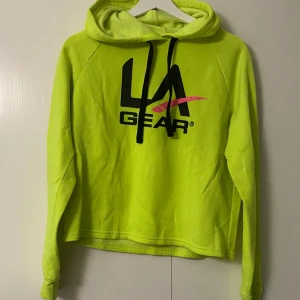 Neongul hoodie från LA Gear - Säljer en cool neongul hoodie från LA Gear. Den har en stor logga på framsidan i svart och rosa. Perfekt för att sticka ut i mängden! Tröjan är i bra skick och har en skön passform med långa ärmar och en klassisk huva. Perfekt för höst och vår.