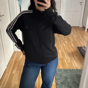 Svart träningsjacka från Adidas - Säljer en svart träningsjacka från Adidas i mycket bra skick. Den har de klassiska tre vita ränderna längs ärmarna och en dragkedja framtill. Perfekt för träning eller som en casual vardagsjacka. Materialet är mjukt och bekvämt, vilket gör den idealisk för både inomhus och utomhusaktiviteter.