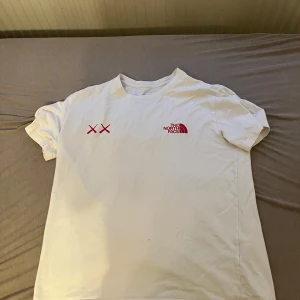 Vit t-shirt från The North Face x kaws - Säljer en snygg vit t-shirt från The North Face i bra skick. Den har två röda kryss som presenterar kaws loggan  och loggan tryckt på bröstet. Perfekt för vardagsbruk eller träning. Passar till allt och är superbekväm!