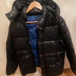 Svart dunjacka från Polo Ralph Lauren - Säljer en svart dunjacka från Polo Ralph Lauren i storlek L. Jackan har en pufferstil med en mysig huva och dragkedja framtill. Perfekt för kalla vinterdagar. Insidan är fodrad med blått tyg för extra värme. Passar både till vardags och mer uppklädda tillfällen.  Storleken är junior  L 14-16 år 