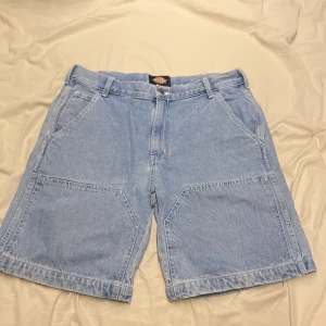 Blå jeansshorts från Dickies - Säljer ett par klassiska blå jeansshorts från Dickies i storlek W34. De har en avslappnad passform med praktiska fickor både fram och bak. Perfekta för sommardagar eller avslappnade tillfällen. Shortsens material är slitstarkt och bekvämt, vilket gör dem till ett måste i garderoben.