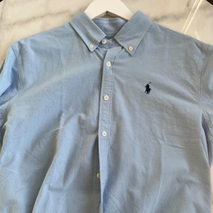 Ljusblå skjorta från Ralph Lauren - Säljer en klassisk ljusblå skjorta från Ralph Lauren i bomull. Skjortan har en broderad logga på bröstet och knappar framtill. Perfekt för både vardag och fest! 💙