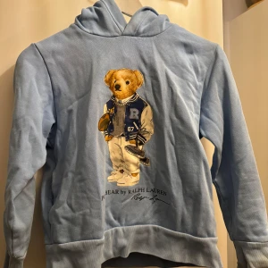 Blå hoodie från Ralph Lauren med björnmotiv - Säljer en superfin ljusblå hoodie från Ralph Lauren med ett gulligt björnmotiv på framsidan. Perfekt för en avslappnad stil och passar bra till både jeans och mjukisbyxor. Hoodien är i bra skick och har en skön passform.  Strl 10-12år