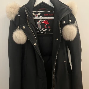 Svart dunjacka från Moose Knuckles - Säljer en snygg och varm svart vinterjacka från Moose Knuckles. Jackan har en fluffig päls på huvan och är perfekt för kalla vinterdagar. Den har en dragkedja framtill och flera knappar. Använd i en vinter. Jackan är i mycket bra skick  7/10. (Pris kan diskuteras)