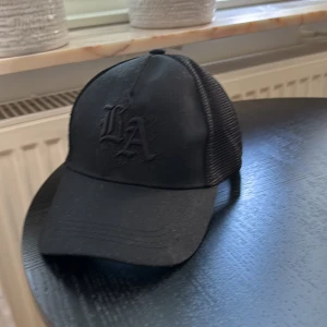 Svart keps med LA-broderi - Säljer en snygg svart keps med LA-broderi framtill. Kepsen har ett nätmaterial baktill för bra ventilation och en justerbar rem för perfekt passform. Perfekt för en avslappnad stil eller en dag på stan!