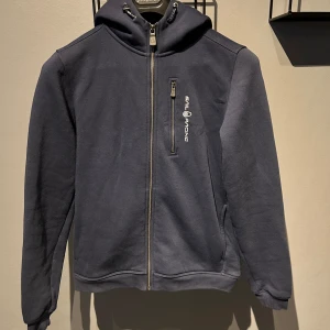 Mörkblå hoodie från Sail Racing - Säljer en snygg mörkblå hoodie från Sail Racing. Den har en dragkedja framtill och en praktisk bröstficka med dragkedja. Hoodien är långärmad och har en bekväm passform. Perfekt för både vardag och fritid!