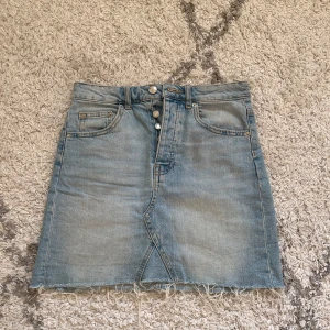 Jeanskjol med rå kant - Säljer en snygg jeanskjol i ljusblå denim med rå kant nertill. Kjolen har en klassisk femficksdesign och stängs med knappar framtill. Perfekt för en avslappnad sommarlook! 🦋