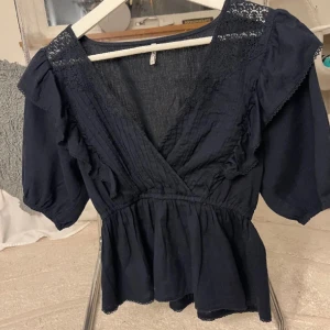 Marinblå blus - Säljer en elegant marinblå blus med v-ringning och spetsdetaljer. Blusen har korta ärmar med volang och en omlott-design som ger en fin passform. Perfekt för både vardag och fest! Köpt för 400kr på only!