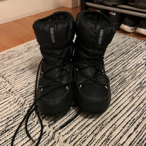 Svarta Moon Boots med snörning - Säljer ett par svarta Moon Boots med snörning. Perfekta för kalla vinterdagar med sin vadderade design och höga skaft. De har en robust sula som ger bra grepp på snö och is. Använda fåtal gånger så nyskick. Storlek 40 men små i storlek, jag har 38 och dom är nästan små för mig. Nypris 2200kr. 