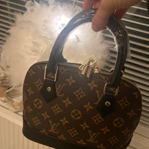 Alma bbBrun handväska med monogram - Snygg brun handväska med klassiskt monogrammönster och svarta handtag. Väskan har en dragkedja upptill och detaljer i guld. Perfekt för att ge en lyxig touch till din outfit.