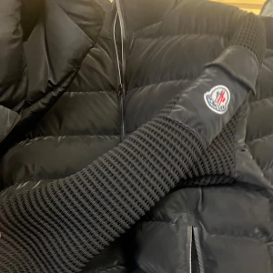 Svart pufferjacka från Moncler - Snygg svart pufferjacka från Moncler med stickade detaljer på ärmarna. Jackan har en dragkedja framtill och två praktiska fickor med dragkedjor. Perfekt för kyligare dagar. priset kan diskuteras det finns många storlekar