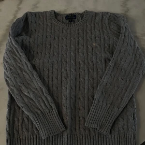 Grå stickad tröja från Ralph Lauren - Säljer en stilren grå stickad tröja från Ralph Lauren med klassiskt flätmönster. Tröjan har långa ärmar och ribbstickade muddar vid ärmslut och nederkant. Perfekt för kyligare dagar!