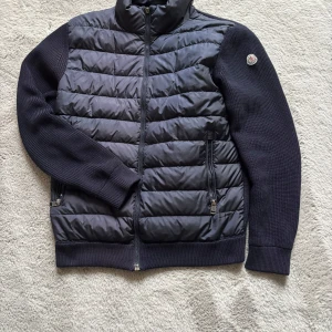 Svart quiltad jacka från Moncler - Snygg svart quiltad jacka från Moncler med dragkedja och ribbade detaljer. Jackan har långa ärmar och en stilren design, perfekt för kyligare dagar. Den man dra i för att zippa up jackan har lossnat men funkar lika bra utan