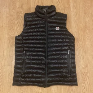 Svart dunväst från Moncler - Snygg svart dunväst från Moncler med dragkedja och hög krage. Perfekt för kyliga dagar. Västen har två praktiska fickor med dragkedja och Moncler-logga på bröstet.