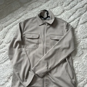Beige/grå overshirt med dragkedja - Snygg beige overshirt med dragkedja och två bröstfickor med lock. Perfekt för lager-på-lager-stil. Långärmad design med klassisk krage.