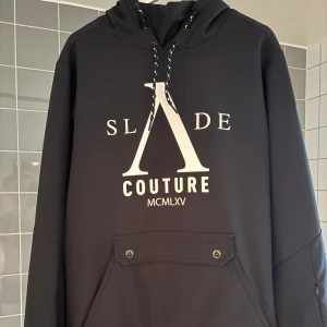 Svart skidjacka från Slade Couture - Snygg svart skidhoodie från Slade Couture med tryck framtill. Jackan är inte använd så mycket så den är i riktigt gott skick!