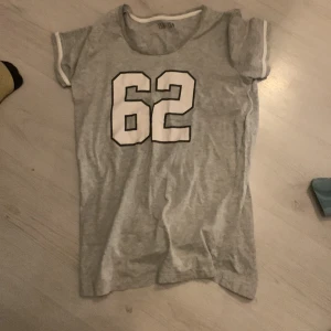 Grå pijamas tröja med nummer 62 - Säljer en grå pijamas tröja med trycket '62' i vitt på framsidan. T-shirten är kortärmad och tillverkad av 95% bomull och 5% viskos, vilket gör den mjuk och bekväm. Perfekt för en avslappnad stil.