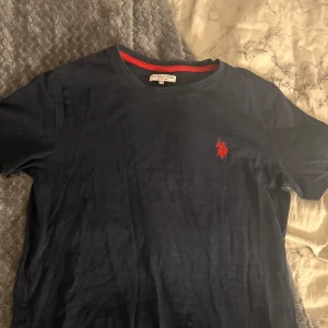 Mörkblå t-shirt från Polo. - Säljer en mörkblå t-shirt från U.S. Polo Assn. med röd logga på bröstet. T-shirten har en rund halsringning med röd kant. Perfekt för en avslappnad stil.