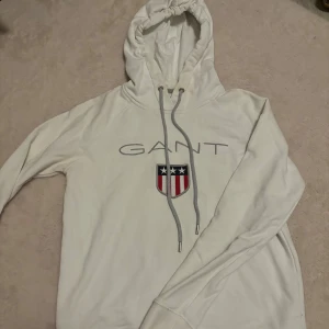 Vit hoodie från GANT - Säljer en stilren vit hoodie från GANT med logotyp och sköldmotiv på bröstet. Tröjan har en justerbar huva med dragsko och långa ärmar. Perfekt för en avslappnad stil.