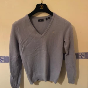 Grå V-ringad tröja från Gant - Säljer en stilren grå V-ringad tröja från Gant. Tröjan är tillverkad i en mjuk ullblandning med 80% ull och 20% polyamid. Perfekt för kyligare dagar med sin klassiska design och diskreta logga på bröstet.