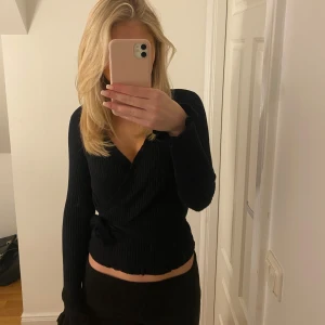 Svart ribbad omlottopp - Snygg svart ribbad omlottopp med lång ärm. Har en figurnära passform och är tillverkad i ett mjukt material. Söta volangärmar. 