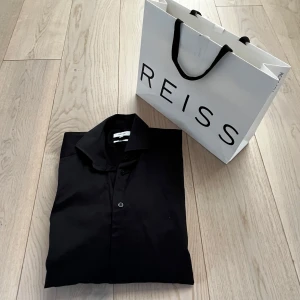 Svart skjorta från Reiss - Snygg svart skjorta från Reiss i slim fit. Perfekt för en stilren look med klassiska knappar och långärmad design. Passar utmärkt för både formella och avslappnade tillfällen. Nypris 1200kr