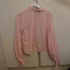 Rosa blus - OANVÄND - En super söt rosa blus med lång ärm och dekorativa band vid ärmsluten. Blusen har en hög krage och en liten öppning i nacken med knappar. Passar perfekt till både vardag och fest. Helt oanvänd!