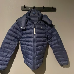 Mörkblå jacka från GANT - Säljer en snygg mörkblå jacka från GANT med dragkedja och avtagbar huva. Jackan har broderad logga på bröstet och är perfekt för kyligare dagar. Passar ungdomar i åldern 13-14 år. Storlek 158-164