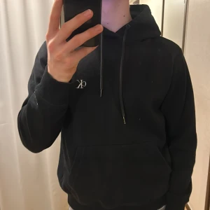 Svart hoodie från Calvin Klein - Säljer en stilren svart hoodie från Calvin Klein med klassisk logga på bröstet. Hoodien har en justerbar dragsko i huvan och en praktisk magficka. Perfekt för en avslappnad look.