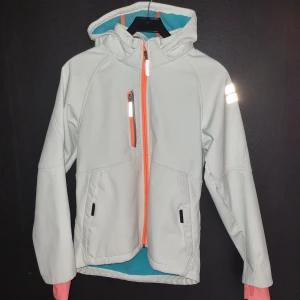 Vit softshelljacka jacka med huva - Snygg vit softshelljacka jacka med huva och orange dragkedja. Jackan har långa ärmar och reflekterande detaljer på ärmen. Perfekt för blåsiga dagar. Den har några fläckar som jag inte har fått bort men de tar inte bort jackans funktion.  Har flera bilder men kunde inte ladda upp fler än 5 st.