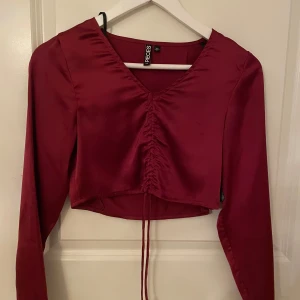 Vinröd topp  - Snygg vinröd topp i ett satin liknade tyg med v-ringning och långa ärmar. Toppen har en trendig snörning framtill och är croppad. Toppen har en smidig dragkedja på sidan. Den passar perfekt både till en elegant outfit eller fest 🎉. I ett fint skick ⭐️storlek xs. 