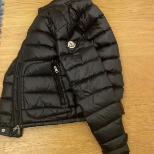 Svart pufferjacka från Moncler - Snygg svart pufferjacka från Moncler med dragkedja och hög krage. Jackan har långa ärmar och två praktiska fickor med dragkedja. Perfekt för kyliga dagar. Säljer den för 1500 eftersom dragkedjan är lite trög. Köpte den från plick för 2000 tusen för ett halvår sen men den var förliten