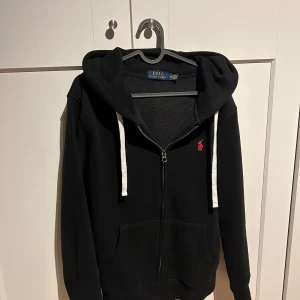 Svart hoodie från Polo Ralph Lauren - Säljer en klassisk svart hoodie från Polo Ralph Lauren med dragkedja och vit dragsko. Den har en broderad röd logotyp på bröstet och är perfekt för en avslappnad stil.