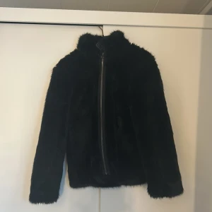 Svart pälsjacka från Lindex - Säljer en svart pälsjacka från Lindex för barn i storlek 152. Jackan har en dragkedja framtill och är långärmad, perfekt för kyligare dagar. Den är mjuk och stilren, vilket gör den till ett mångsidigt plagg i garderoben.