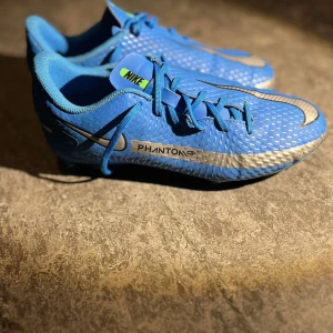 Blå Nike Phantom fotbollsskor - Snygga blå Nike Phantom fotbollsskor med silverdetaljer och grön sula. Skorna har snörning och är designade för optimal prestanda på planen. Perfekta för fotbollsspelare som vill ha stil och funktionalitet.