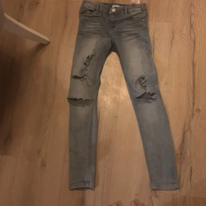 Blå jeans från Name It - Säljer ett par blå jeans från Name It med slitna detaljer på knäna. De har en skinny passform och är perfekta för en avslappnad stil. Jeansen har klassisk femficksdesign och knappgylf.