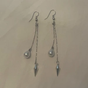 Silverfärgade örhängen  - Eleganta silverfärgade örhängen med hängande kedjor. Ena kedjan har en vit pärla och den andra en spetsig detalj. Perfekta för att ge en stilren touch till din outfit. Rostfritt stål