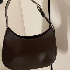 Väska Wera Åhléns  - Snygg mörkbrun axelväska från Wera i skinn med justerbart spänne och dragkedja. Rymmer mycket! Köpt på Åhléns för 959kr. Inga märken eller fläckar. Mindre fack i väskan